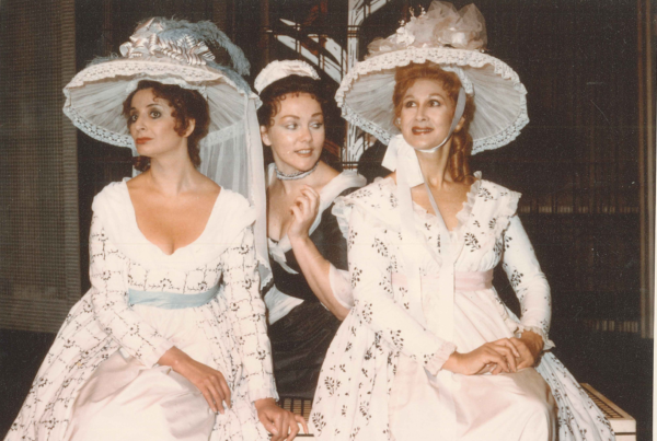 The three women sit in costume; two in lace white dresses with complimenting hats, and one woman dressed as a maid